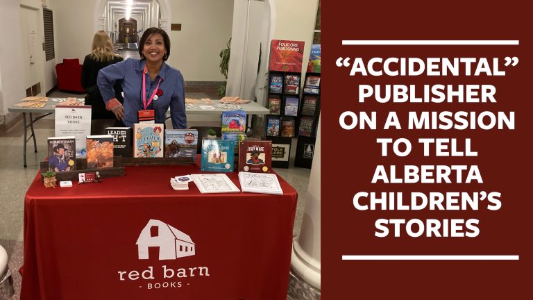 In white text against a red background is the following, “Accidental Publisher On a Mission to Tell Children’s Stories”. Beside the text is a photograph of Ayesha Clough, of Red Barn Books, standing behind a table displaying her recently published books.