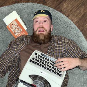 Colby Clair Stolson lies on a rug with a stunned expression on his face. A typewriter rests on his stomach, and books are on the ground beside him. 
